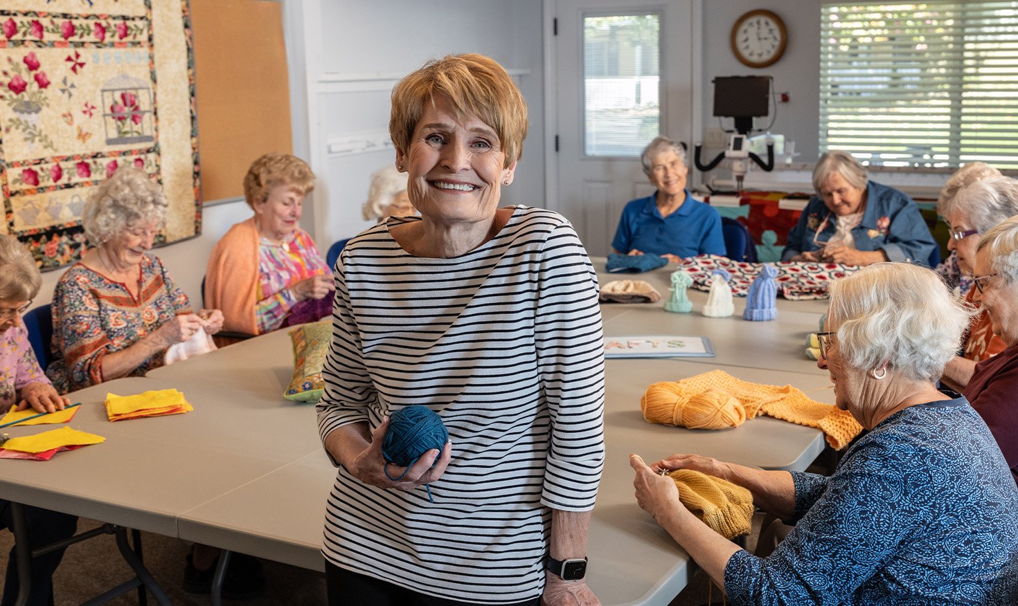 Senior woman holding a ball of yarn at the head of a table full of women working on knitting projects