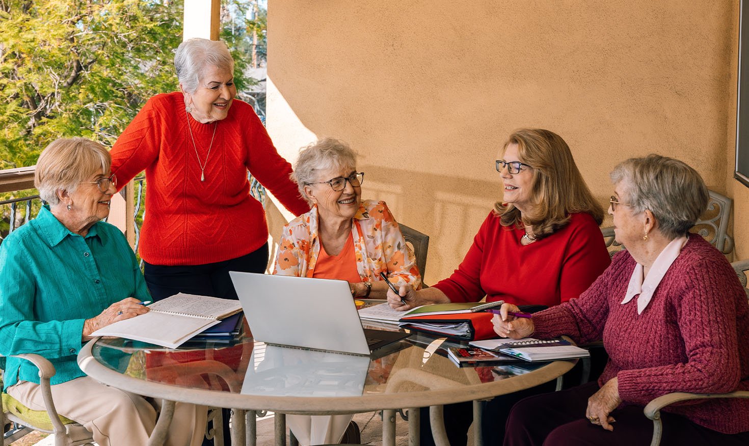 Four senior women sitting and one standing with notebooks sit around a table during a writers' group meeting at Plymouth Village