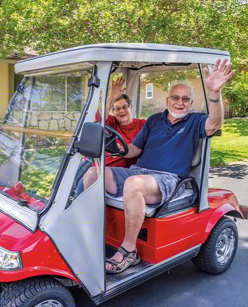 Senior man and senior woman waving from a red golf cart while driving around Plymouth Village