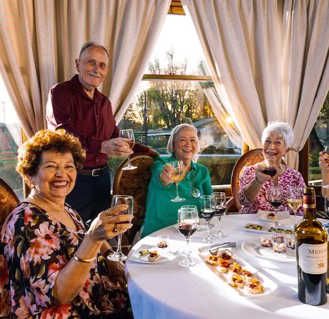 Four senior friends in the Plymouth Village dining room holding up glasses of wine