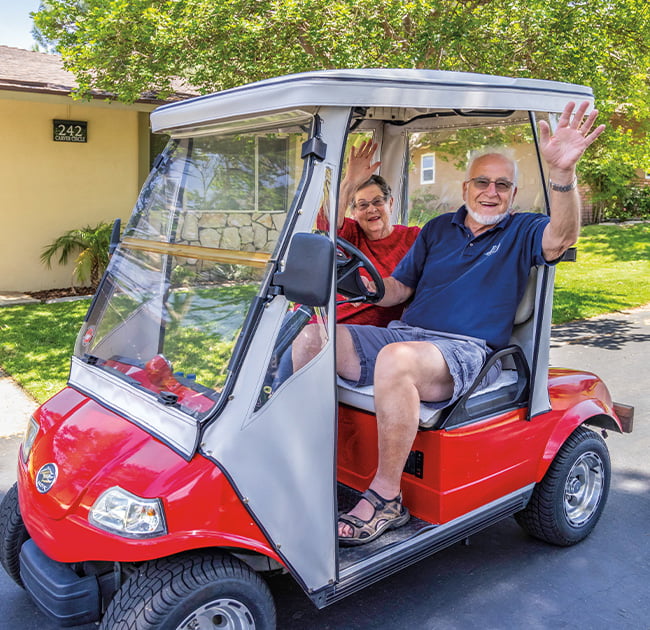 Senior man and senior woman waving from a red golf cart while driving around Plymouth Village