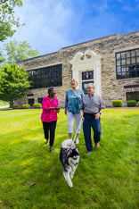 Two senior women and a senior man walking a husky dog near the exterior entrance of the Spring Mill Pointe community