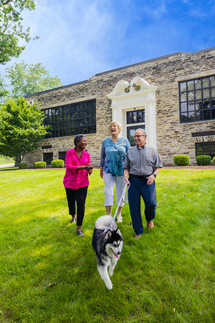 Two senior women and a senior man walking a husky dog near the exterior entrance of the Spring Mill Pointe community