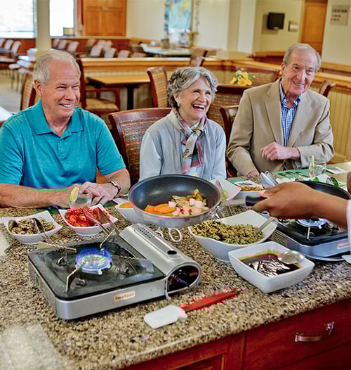 Two senior men and a senior woman enjoying a meal that is freshly prepared in front of them by a chef
