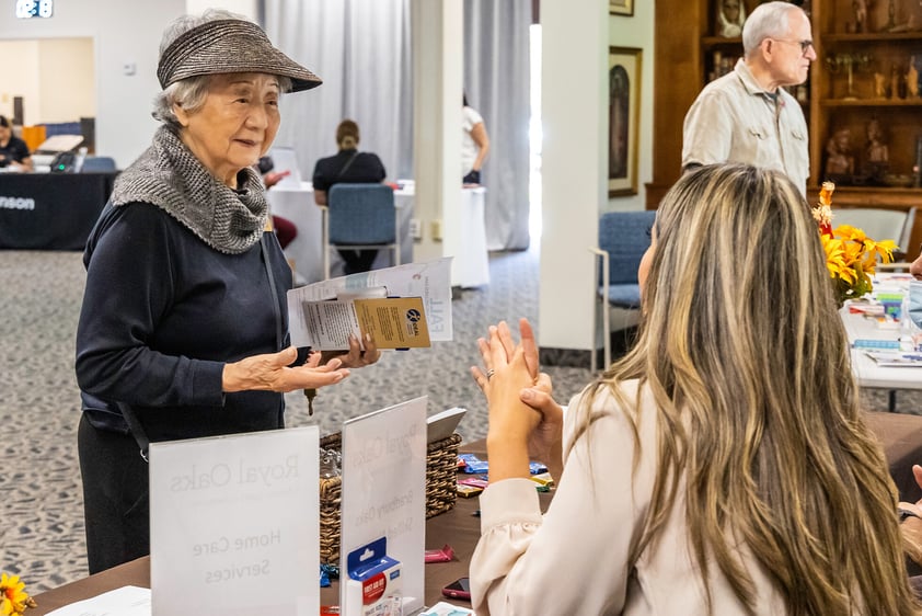 Senior woman holding brochures and talking to a woman at a table during an expo at Westminster Gardens