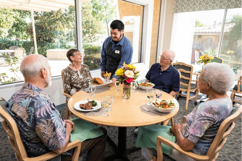 Dining room team member serving food to a group of four seniors in the Westminster Gardens dining room