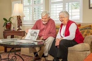 Senior couple sitting on their couch in their Westminster Gardens home looking at sketchbook 