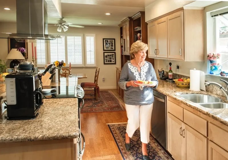 Senior woman holding a plate of food and looking out the kitchen window at her Westminster Gardens home