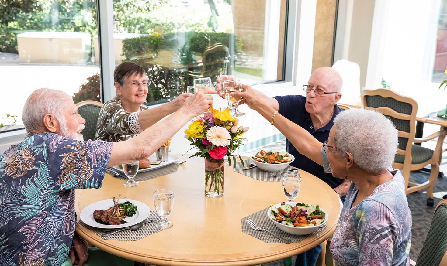 Two senior men and two senior women toasting with glasses of white wine during a meal in the Westminster Gardens dining room