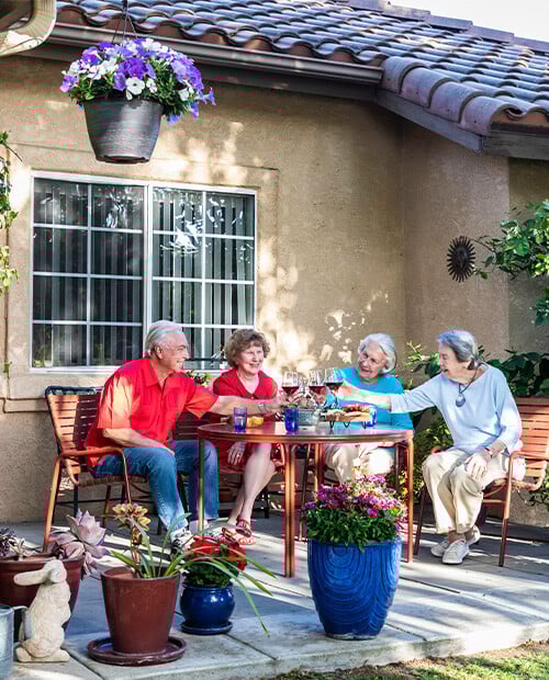 Three senior women and a senior man sitting around a table and toasting with glasses of wine on the patio of a Westminster Gardens home