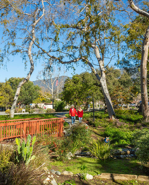 Senior couple wearing red jackets walking toward a bridge on the landscaped grounds of Westminster Gardens
