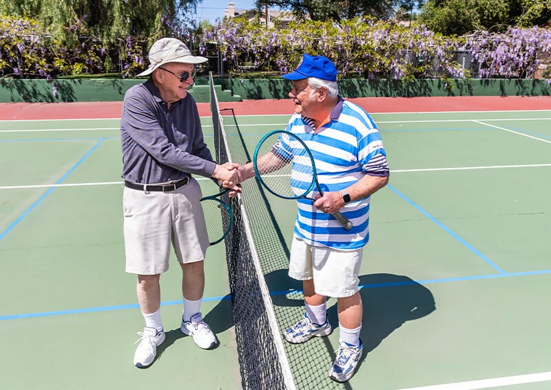 Two senior men holding tennis rackets shaking hands over the net on the Westminster Gardens tennis court 