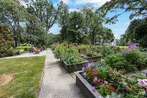 A garden at the Mansion at Rosemont with a central pathway, colorful flowers in raised beds on the right, and chairs on a patio to the left
