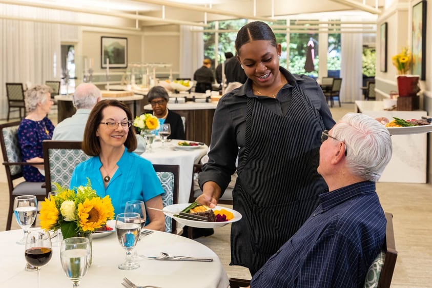 Dining room worker delivering a plate of food to a senior man who is dining with a senior woman in the dining room at Rosewood