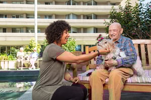Woman petting a dog that's sitting on a senior man's lap on a bench outside Piedmont Gardens