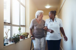A nurse walks a woman with a cane down a hall. 