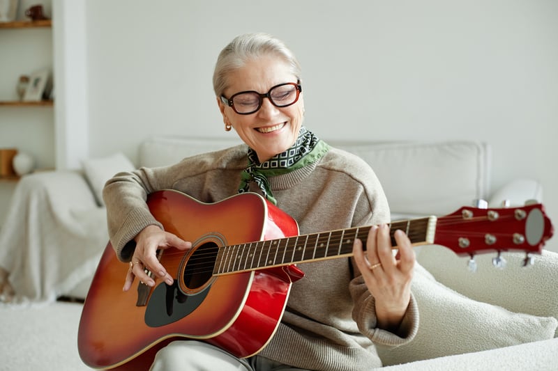 senior woman wearing glasses, siting down and playing guitar.