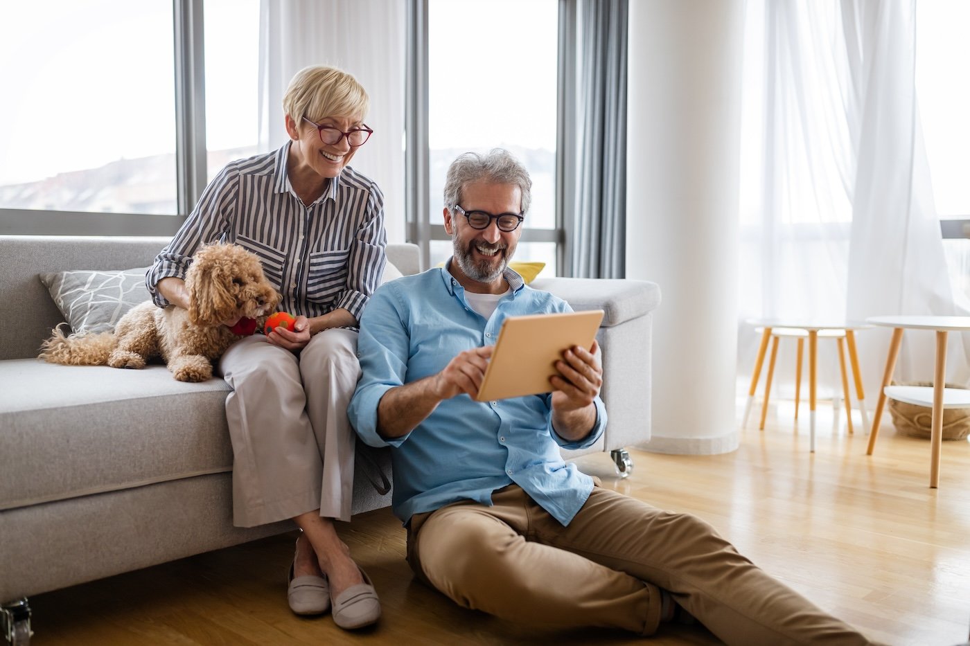 man and woman smiling on couch with their dog looking at a tablet