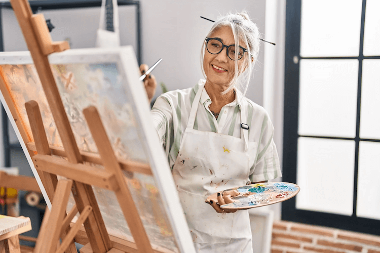 older woman in white apron painting on a canvas while holding a paint palette