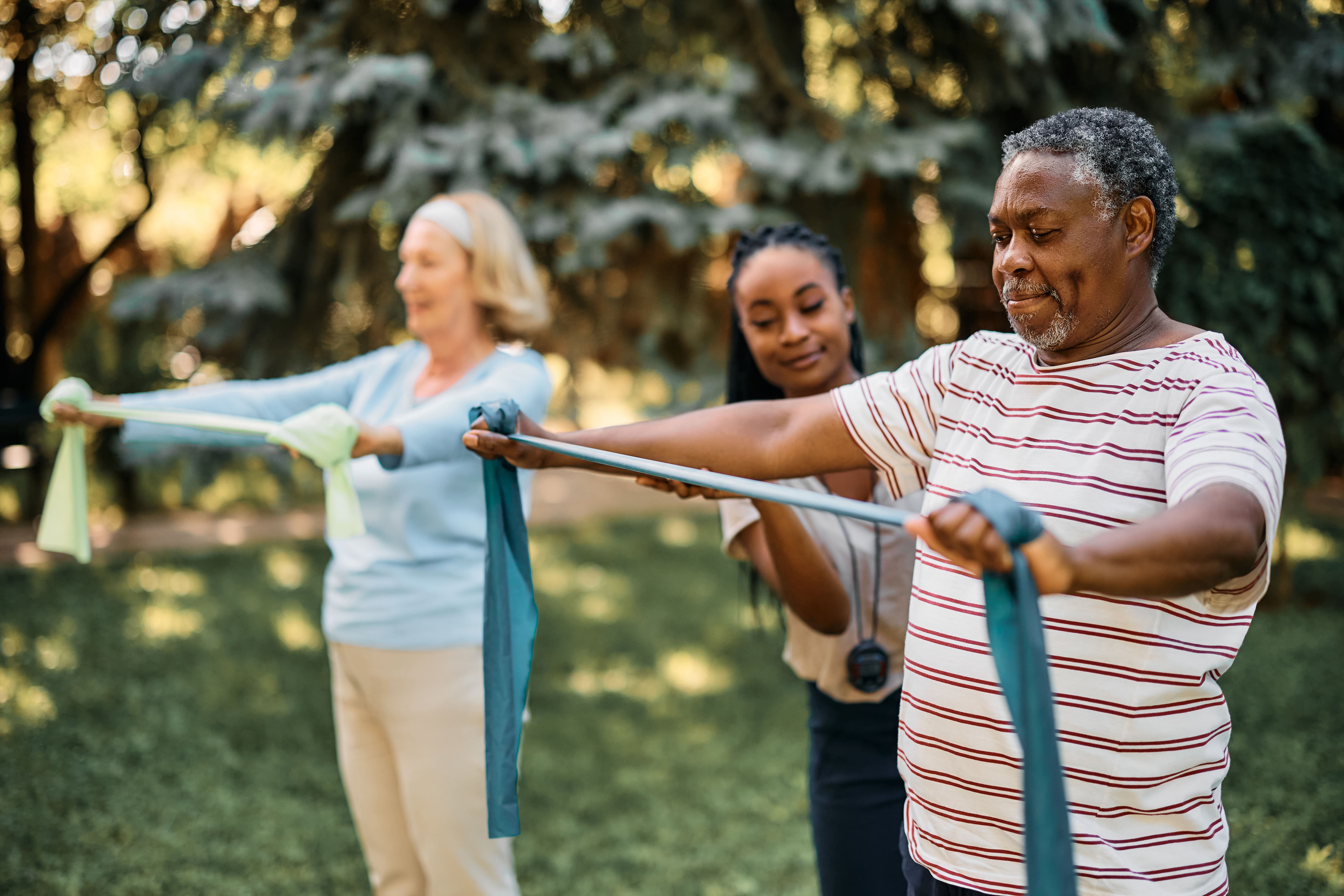 older adult man and woman using exercise bands outside with a trainer