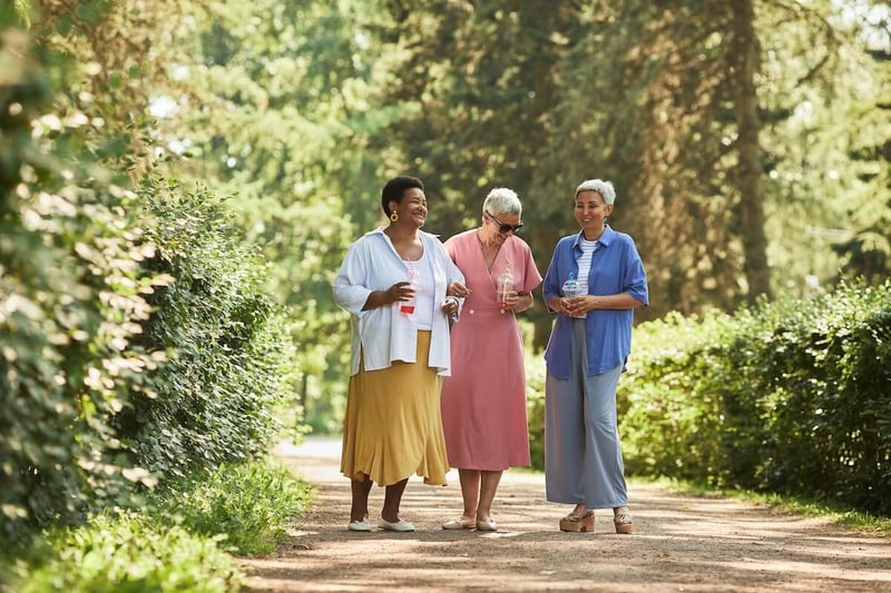 group of 3 senior women walking through a wooded path holding drinks and smiling