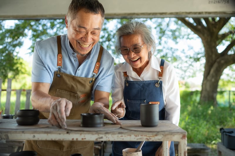 Couple joyfully making pottery outdoors, wearing aprons. 