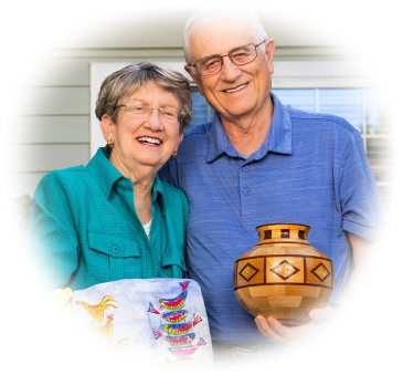 A couple smiles outdoors, holding a decorative vase and a colorful embroidered cloth.