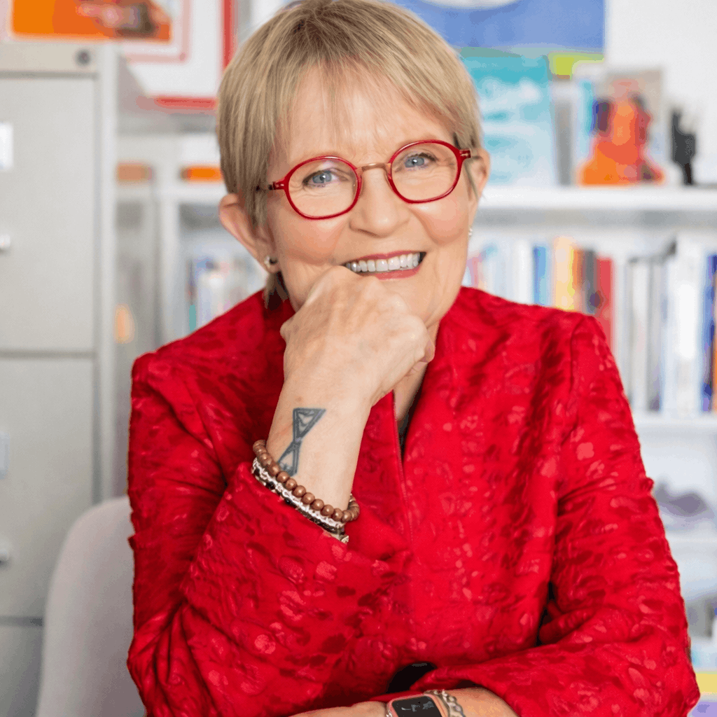 Smiling resident named Sara wearing red glasses and a red jacket, seated indoors.