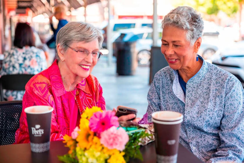 Two senior women enjoying coffee and looking at a cellphone on a patio