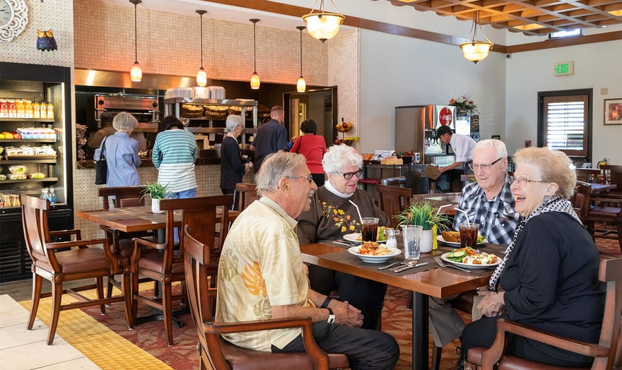 Two senior women and two senior men eating a meal in the community bistro with a line of seniors getting food in the background