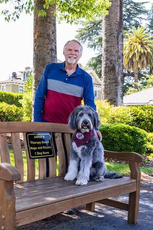 Senior man standing behind an outdoor bench with a dog sitting on it at Plymouth Village