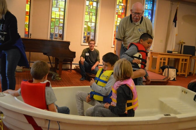 Kids wearing life jackets sit in a boat indoors.