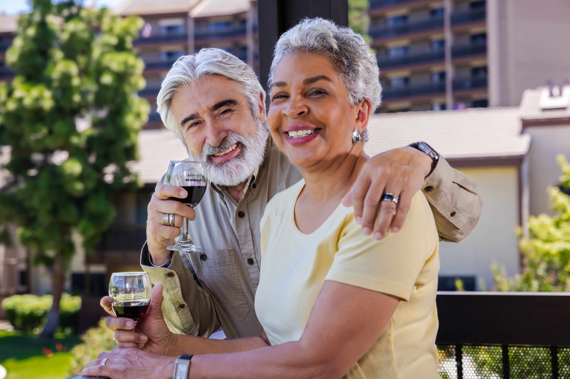 Two residents smile on a balcony at Rosewood, holding glasses of red wine. Two residents smile on a balcony at Rosewood, holding glasses of red wine.