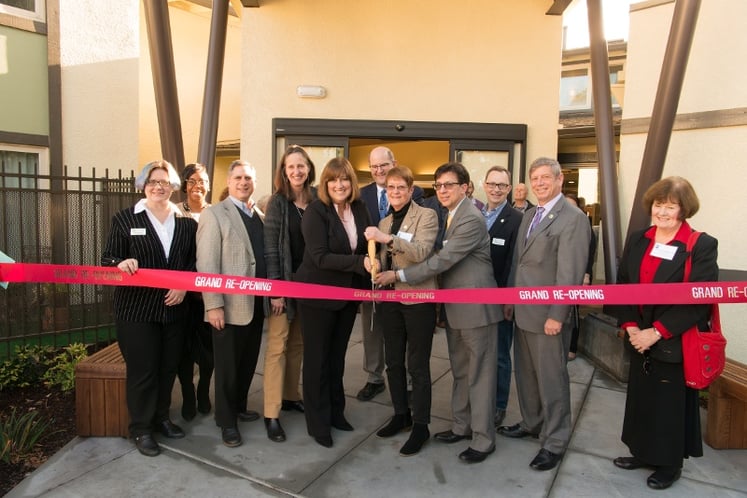 Group cutting a ribbon with big scissors. The ribbon reads 