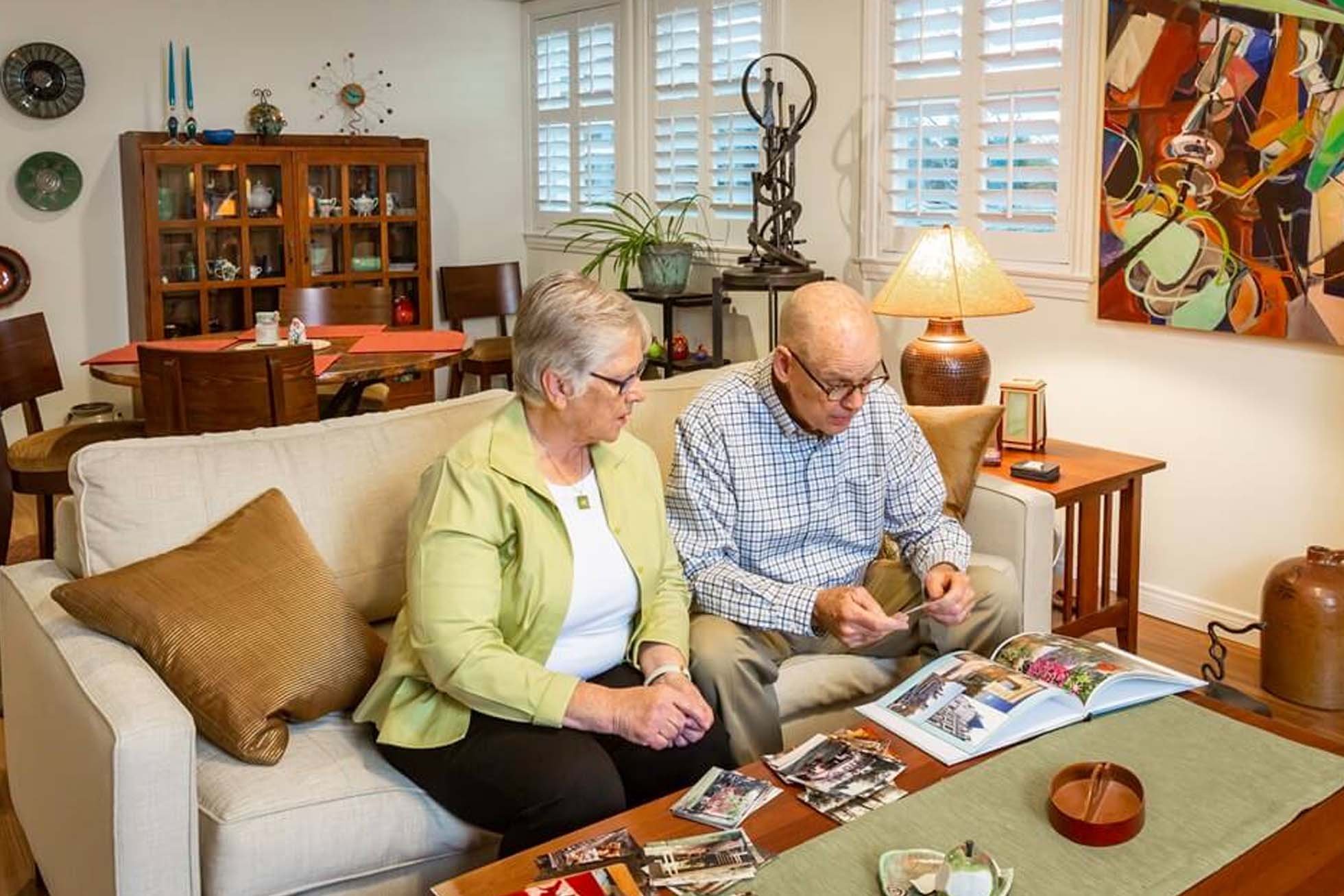 Couple sits on a beige sofa in a cozy living room, looking at photo albums and prints.