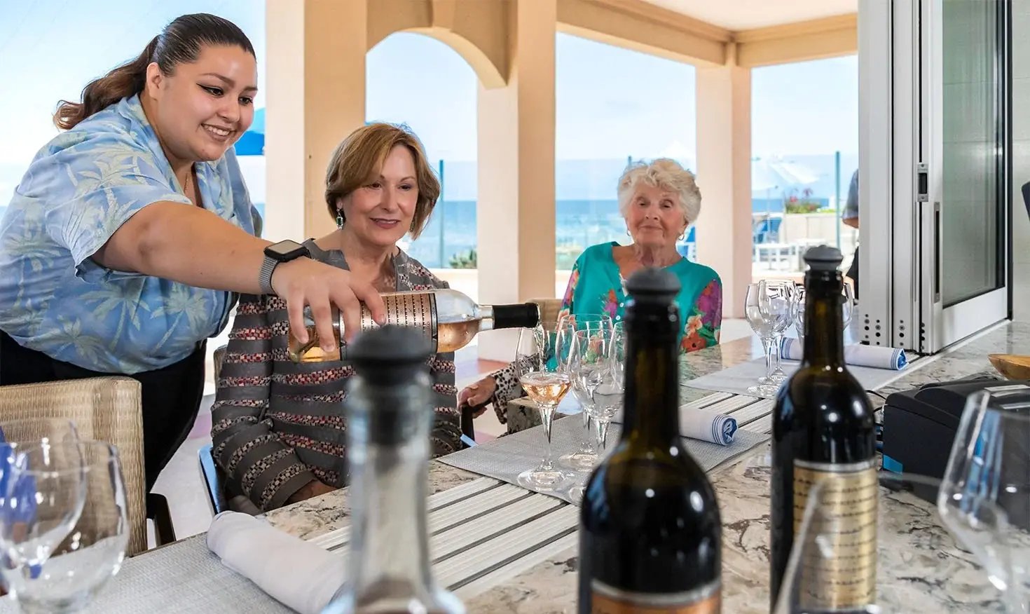 Server pouring wine for two older women at a seaside restaurant.