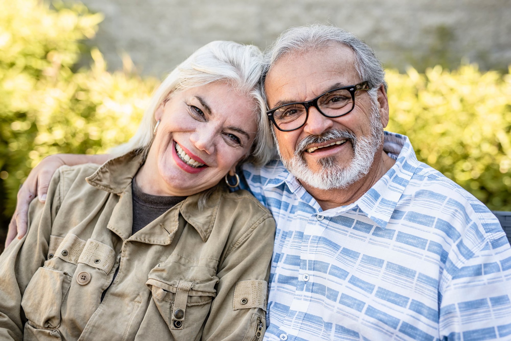 A smiling couple outside on a sunny day with green plants in the background. A smiling couple outside on a sunny day with green plants in the background.