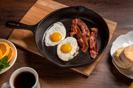 A cast-iron skillet with sunny-side up eggs and bacon sits on a cutting board on a table with biscuits, orange slices, and coffee.