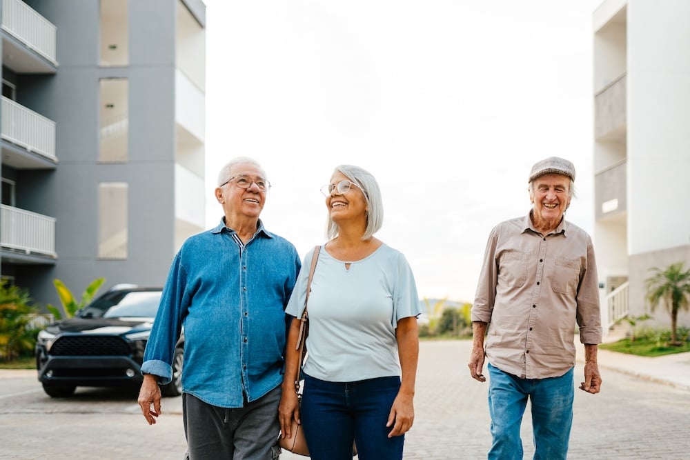 Three smiling adults walk together outdoors, surrounded by modern buildings and a parked car. 
