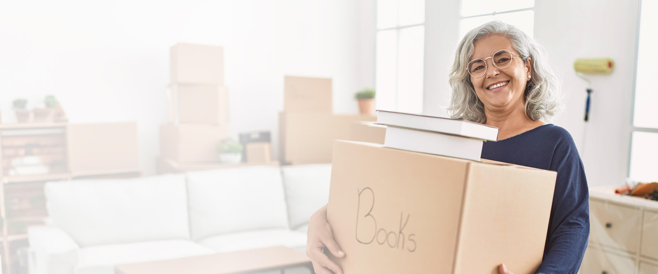 Senior woman holding a moving box with the word "Books" written in marker on the front standing in her home