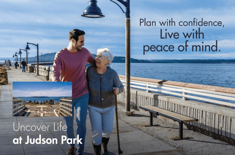 A man and a resident walk arm-in-arm along a waterfront promenade, smiling.