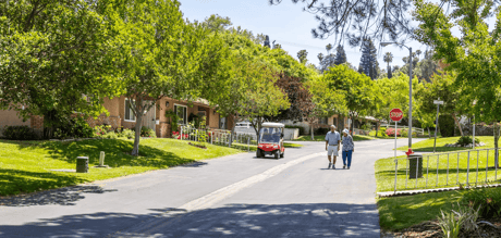 A tree-lined residential street at Plymouth Village with two adults walking and a small red golf cart driving nearby.