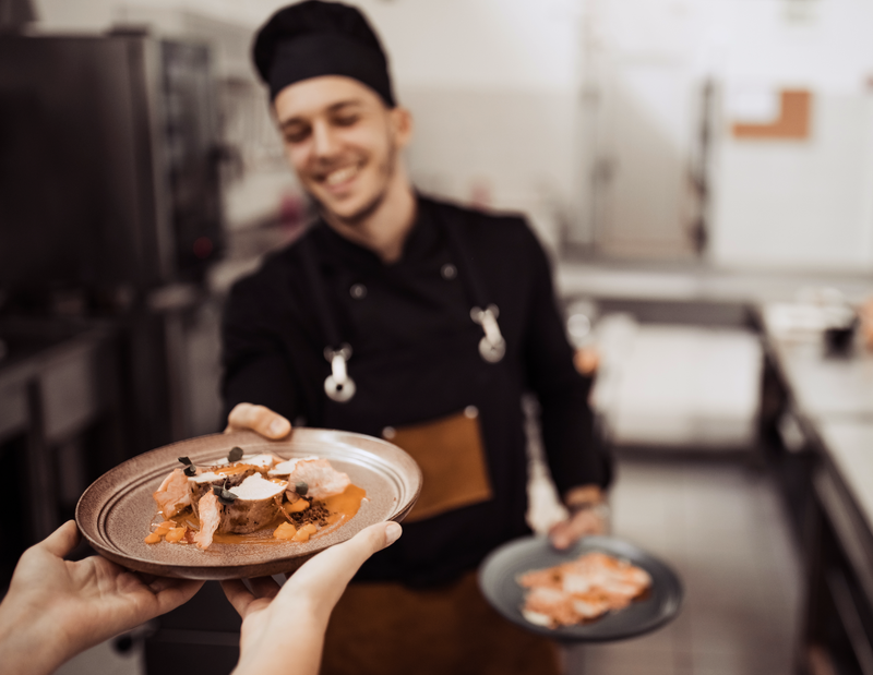 A smiling chef in a black uniform hands over a plated gourmet dish to a resident in a kitchen setting. A smiling chef in a black uniform hands over a plated gourmet dish to a resident in a kitchen setting.