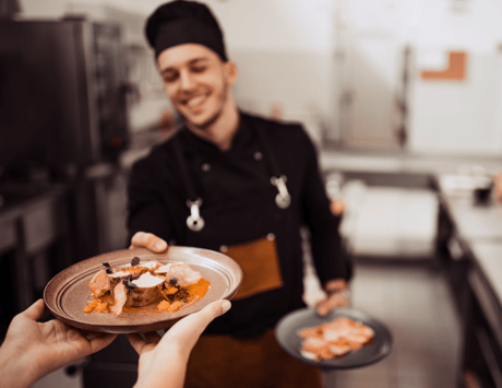 A smiling chef in a black uniform hands over a plated gourmet dish to a resident in a kitchen setting.
