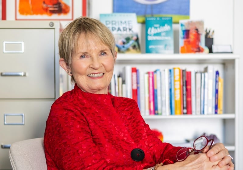 Dr. Sara Hart in a red jacket sits smiling on a beige chair, holding glasses.