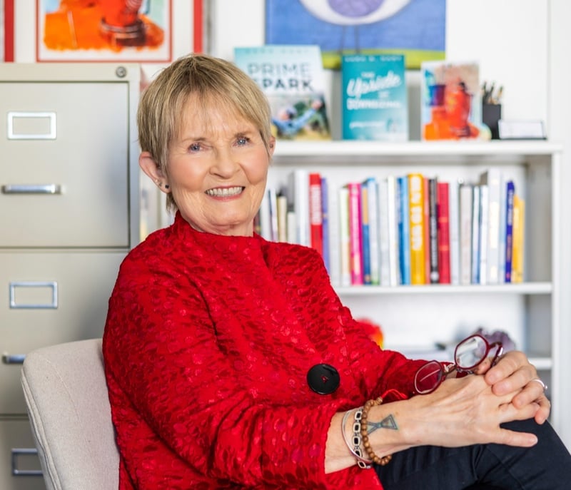 Dr. Sara Hart in a red jacket sits smiling on a beige chair, holding glasses.