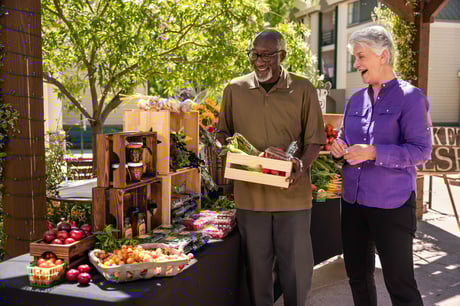 Two smiling residents browse produce at a farmer’s market stall. 