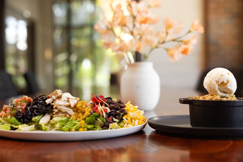 A plated salad and a dessert with ice cream in an elegant dining setting with flowers in the background.