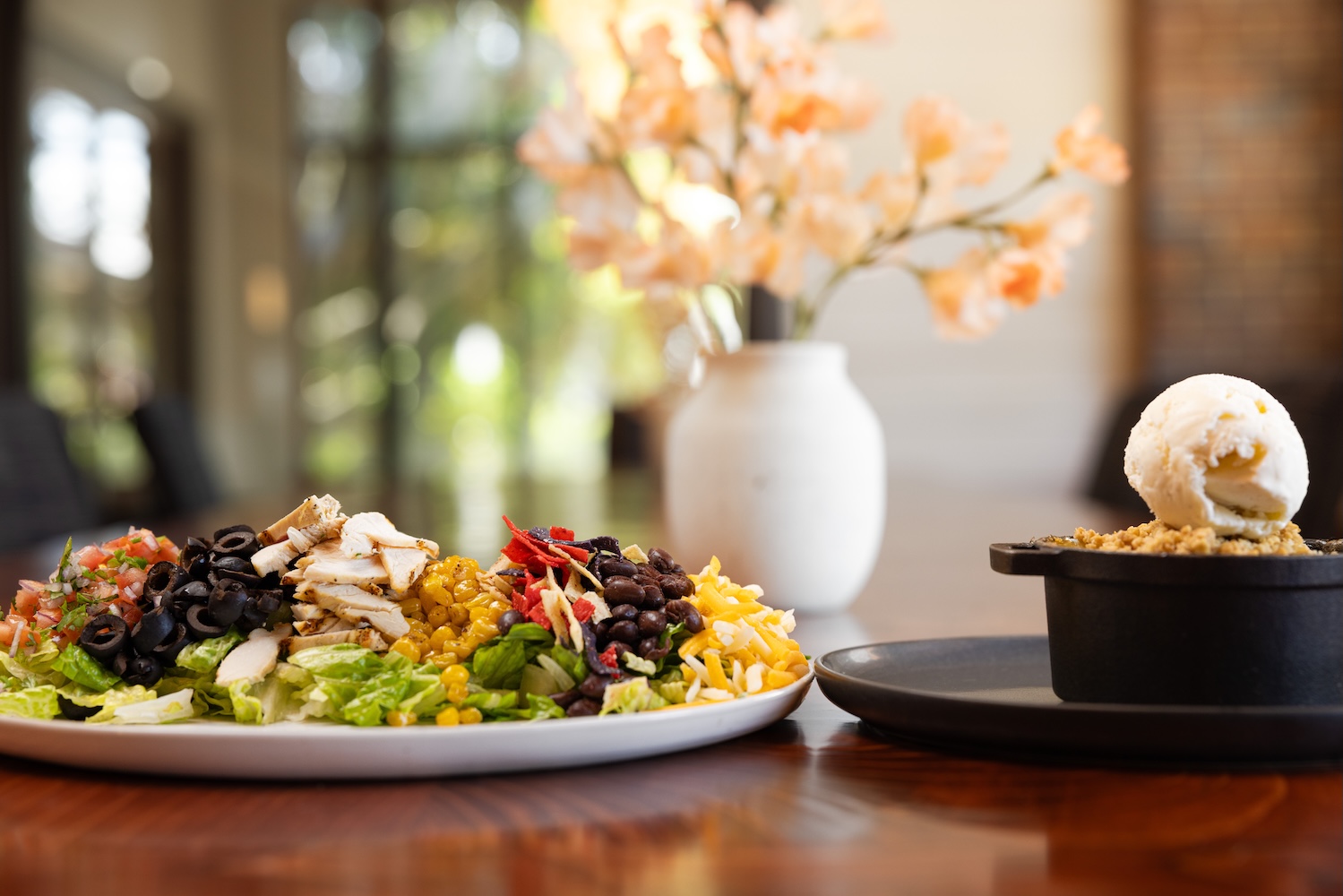 A plated salad and a dessert with ice cream in an elegant dining setting with flowers in the background.