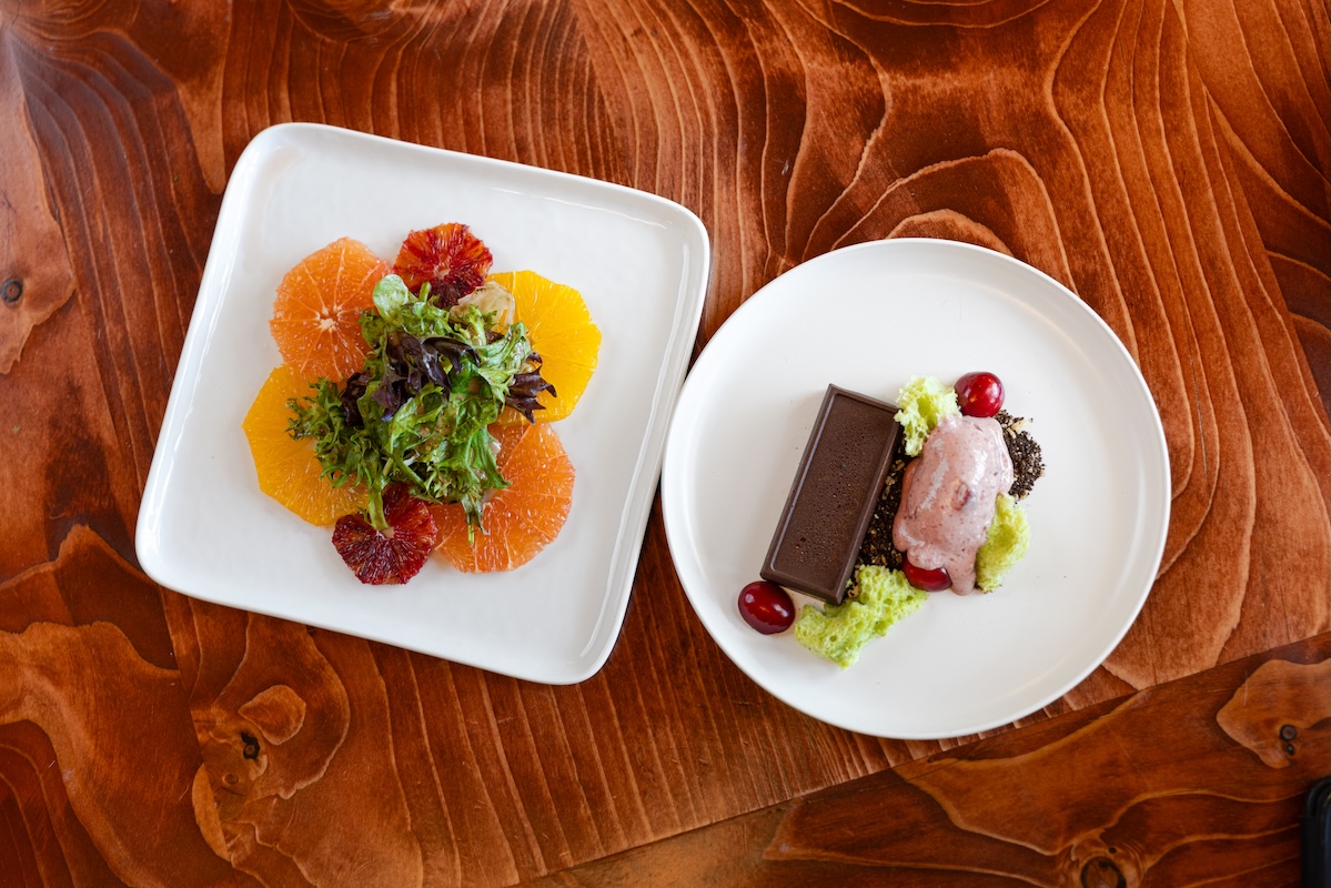 Two white plates with a citrus salad and a chocolate dessert on a wooden table.